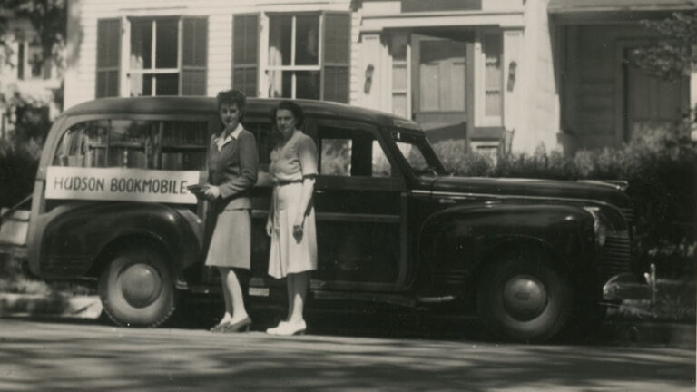 Head Librarian Marjorie Clelland (left) and Natalie Limbach (right) standing in front of bookmobile car in front of the old Hudson Library & Historical Society. Car has a sign that reads, "Hudson Bookmobile"